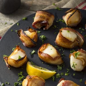 Prosciutto wrapped scallops on black plate with lemon wedge and parsley.